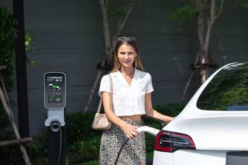 Young woman travel with EV electric car charging in green sustainable city outdoor garden in...