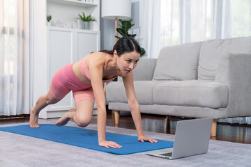 Asian woman in sportswear doing burpee on exercising mat as home workout training routine. Attractive girl engage in her pursuit of healthy lifestyle with online exercise training video. Vigorous