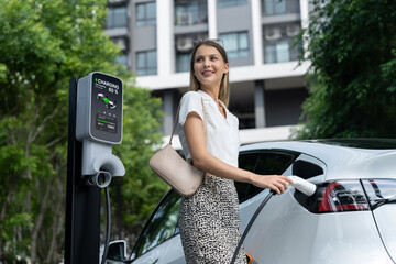 Young woman travel with EV electric car charging in green sustainable city outdoor garden in...