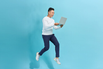 Cheerful asian young man holding laptop smiling jumping high running isolated on blue background. - 654637426