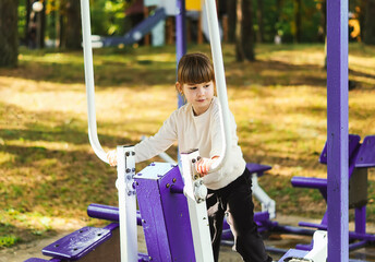 A cheerful five-year-old girl is training on a simulator. Cute little girl on the playground emotionally doing sports. Cute little preschooler training on sports equipment. Healthy childhood concept