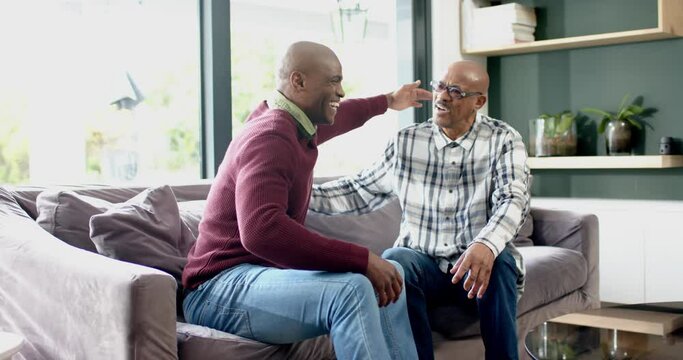 Happy african american senior father and son sitting on couch,embracing and laughing, slow motion