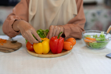 A young Muslim woman in a hijab prepares colorful vegetables on a wooden plate. Useful for making healthy food menus, vegetable salads, while preparing for online blogger broadcasts in the kitchen.