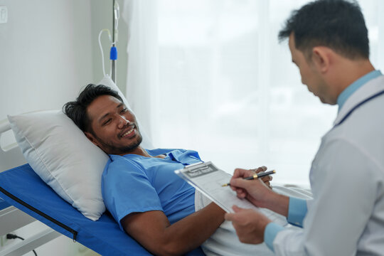 A young Asian doctor and a male patient lying on a hospital bed are discussing medical history and using a clipboard.