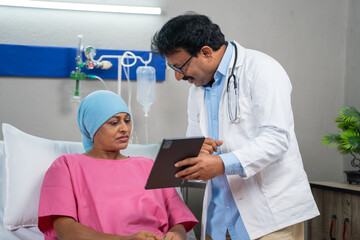 Indian doctor explaining health report from digital tablet to admitted cancer patient at hospital -...
