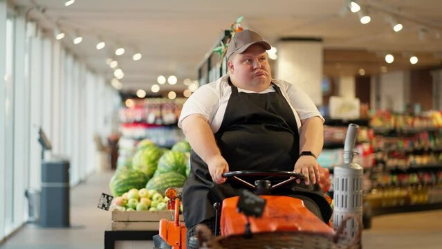 A happy overweight supermarket worker in a white T-shirt and a black apron sits on a special cleaning machine in a cap and grimaces. A modern cleaner uses a machine to clean the floor in a supermarket