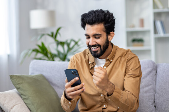 Young man received an online notification of a winning message sitting at home in the living room on the sofa, the winner is satisfied with results of the achievement joyfully holding his hand up
