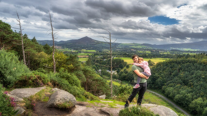 Father holding daughter in arms and standing on edge of cliff, with a scenic view on a valley and...