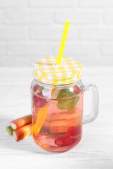 Mason jar of tasty rhubarb cocktail with raspberry and stalks on white wooden table, closeup