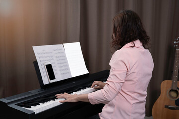 An asian woman in pink shirt and short spiral hair playing paino at home, smartphone and key note...