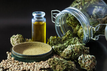 noir still life of hemp products consist of herb grinder full of cannabis pollen surrounded by dry flowers of medical marijuana, glass jar with buds and small bottle with CBD oil close up on black bac