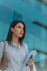 Gorgeous brunette businesswoman holding paperwork and smart phone looking away while standing in front of big business center