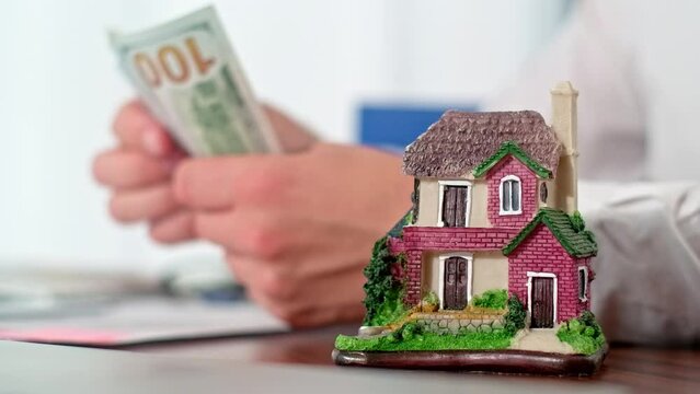 Man counting paper dollar currency with house figurine in front. Concept of renting a house or paying tax for an apartment. First payment of a mortgage loan