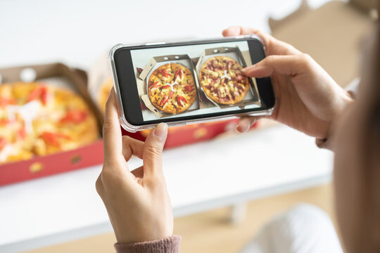 Happy meal, enjoy eating beautiful asian young woman taking photo of pizza in box by mobile smart phone on table at home before eat fast or junk food, post picture to communication on social networks.