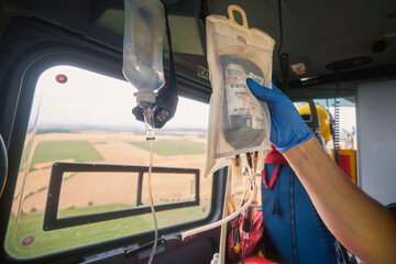 Hand of doctor holding transfusion bag with blood on board helicopter of emergency medical service during flight to hospital. Themes rescue, urgency and health care.. - obrazy, fototapety, plakaty