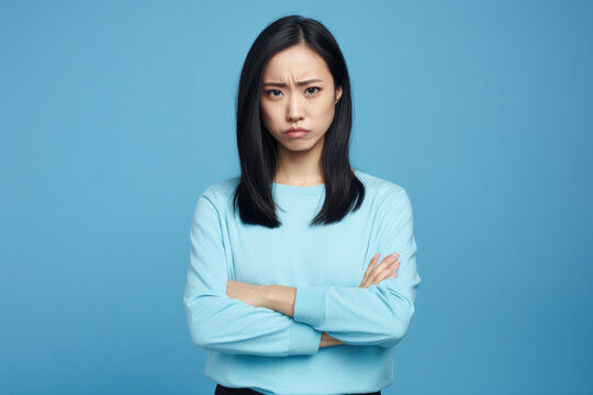 Offended Asian Woman: Portrait of a Dissatisfied Young Lady with Folded Arms, Expressing Disagreement, Isolated on Blue Studio Background.