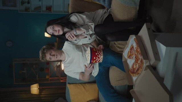 Boy and girl sitting on the sofa eating popcorn, watching movie. Boy shyly puts hand on girl's shoulder, girl accepts the hug. Vertical video.