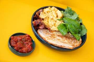 protein breakfast dish with tomato with eggs bacon arugula and slice of sourdough bread on yellow background