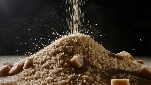 Loose and lump sugar. Brown cane sugar is poured onto table in heap. Close up slow motion. Being wary of your sugar intake is imperative in battle against obesity, diabetes, and other serious ailments