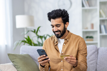 Young joyful man sitting on sofa at home, using application on phone for online shopping in online...