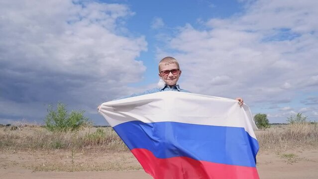 Portrait of a cute, smiling happy boy in a field with a Russian flag on his shoulders. National holiday independence day. Pride and patriotism