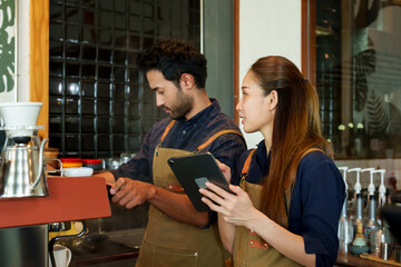 Young barista Coffee shop owner is preparing to make coffee. According to orders that customers order through online applications on tablets. beautiful asian woman and husband Check coffee orders .