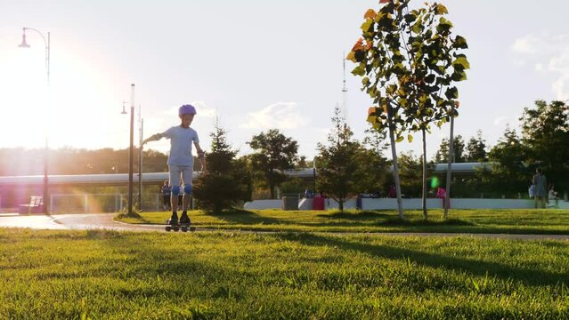 A happy kid in a safety helmet and suit skateboarding in the park. A boy skateboarding on the street in the sunset. Active recreation of children. a family, a child in a park with green grass.