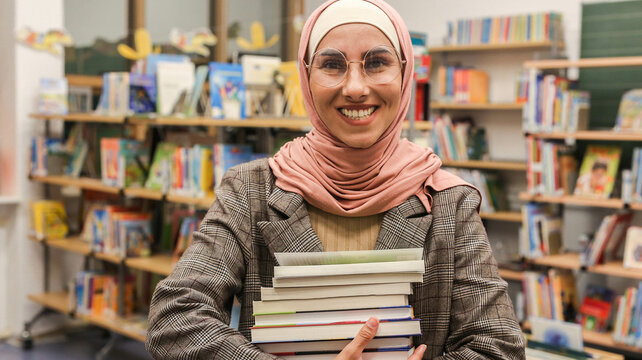 Smiling pretty Muslim Middle Eastern woman in hijab and jacket with glasses holds many books in her hands received textbooks, bought in bookstore. Librarian secretary trainee assistant manager student
