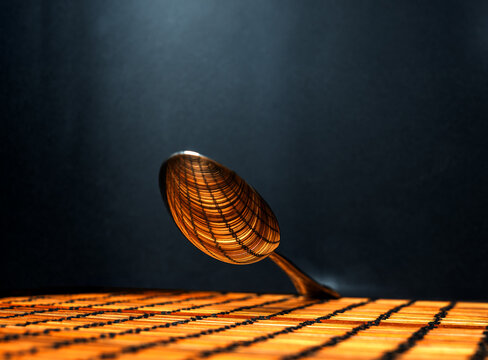 shot of the convex part of a suspended silver spoon reflecting the linear design of the underlying plane, illuminated by a beam of light from above, against a black background