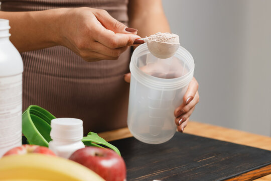 Athletic woman in sportswear with measuring spoon in her hand puts portion of whey protein powder into a shaker on wooden table with amino acid white capsules, bananas and apple, making protein drink