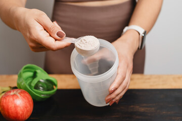 Athletic woman in sportswear with measuring spoon in her hand puts portion of whey protein powder...