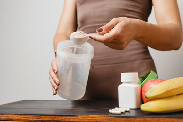 Athletic woman in sportswear with measuring spoon in her hand puts portion of whey protein powder into a shaker on wooden table with amino acid white capsules, bananas and apple, making protein drink