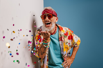 Happy senior man in funky shirt and bandana blowing colorful confetti on white and blue background
