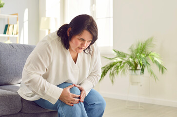 Overweight woman suffering from knee pain. Young fat girl with leg injury sitting on sofa at home...