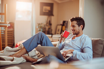 Young man having coffee while using the laptop on the couch at home