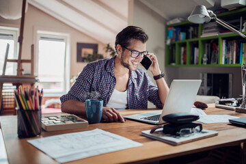 Fototapeta premium Young male architect having a conversation on the smartphone while working on design from home