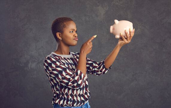 Funny frowning dark-skinned woman waves a punishing finger at the piggy bank making a gesture of shame. Concept of saving money, setting a financial goal and planning future expenses. Gray background.