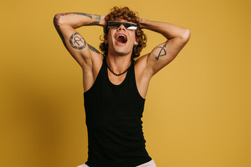 Excited young man in trendy glasses holding hands in hair and smiling against yellow background