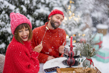 Man and woman in red winter clothes have fun while eating fondue, sitting together by the table...