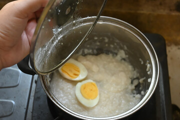 A woman's hand is opening the lid of a pot containing rice porridge and boiled eggs as a baby's breakfast menu.