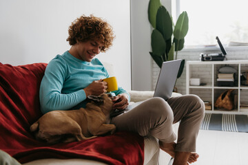 Handsome young curly man petting his dog while working on laptop from home