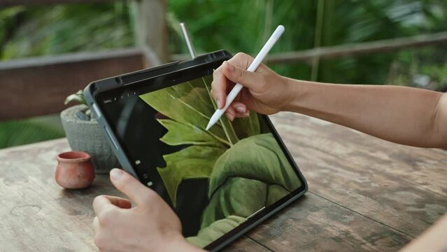 Female artist hands drawing with stylus on tablet at the cafe on tropical island, palm trees. Young designer works remotely open air at exotic location. Pretty woman editing illustration. Close-up.