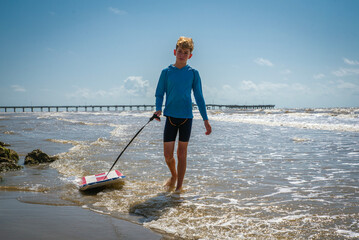 Male child on beach pulling his boogie board while walking and splashing in the surf