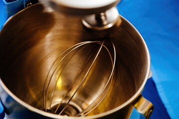 A clean mixer bowl and its metallic attachments sit against a vibrant blue background, ready for...