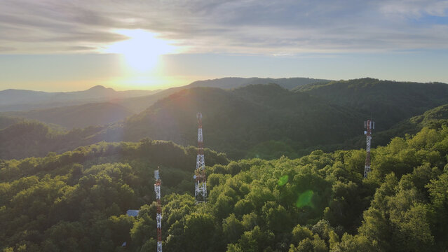 Aerial view of 5G telecommunication towers standing tall on dense green mountains against beautiful sunlight