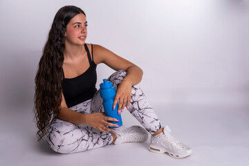 "Young Caucasian woman sitting and resting while drinking water from her bottle against a white background."