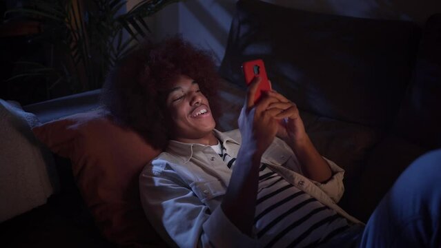 Smiling Latin man looking at cell phone relaxing in living room at home. Happy young male using or chatting mobile lying on sofa at night. Generation z people, social networks and dating apps indoors.