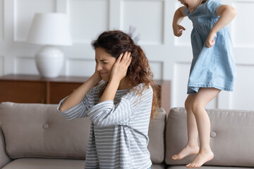 Tired angry young mother sit on couch close ears bothered by ill-behaved naughty little daughter...