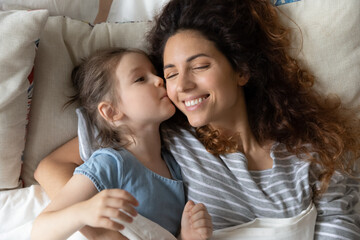 Cute little preschooler daughter and happy millennial mom waking up in cozy home bed together, smiling young mother or nanny relaxing in comfortable bedroom with small girl child cuddling and hugging - obrazy, fototapety, plakaty