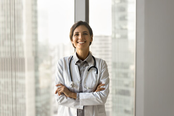 Happy beautiful Latin doctor woman posing at large office window, standing with arms folded, looking at camera with toothy smile. Positive confident medical professional with stethoscope portrait